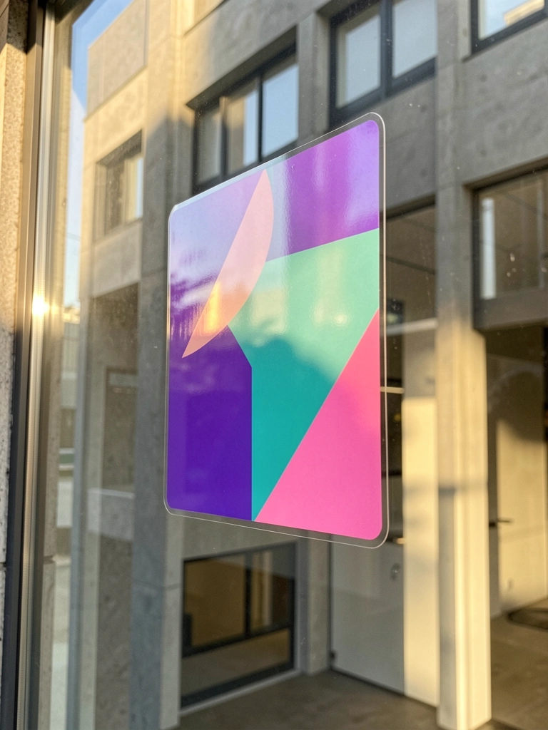 Adesivo transparente com formas geométricas roxas, azuis e rosas em vitrine de loja, refletindo prédio moderno e luz natural do dia.