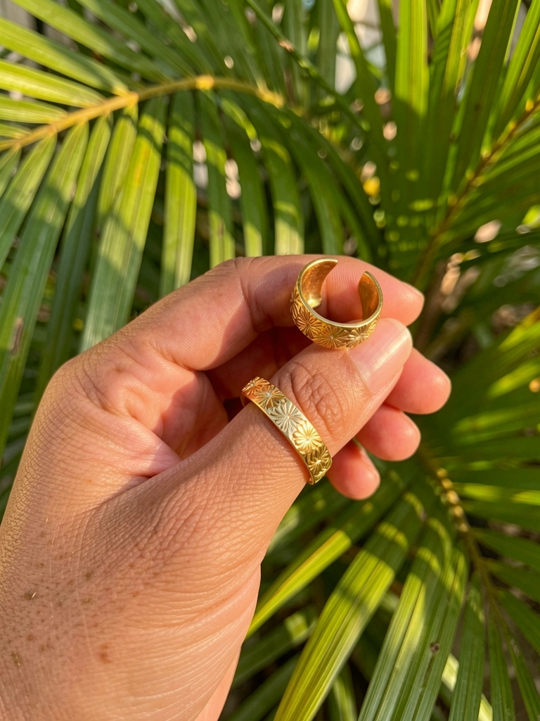 Mão feminina segurando dois anéis de ouro com detalhes florais, palmeiras verdes desfocadas ao fundo com luz tropical.
