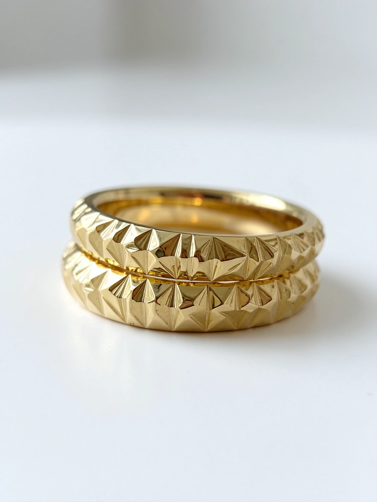 Dois anéis de ouro amarelo com textura facetada e brilho suave sobre superfície branca, um dentro do outro.