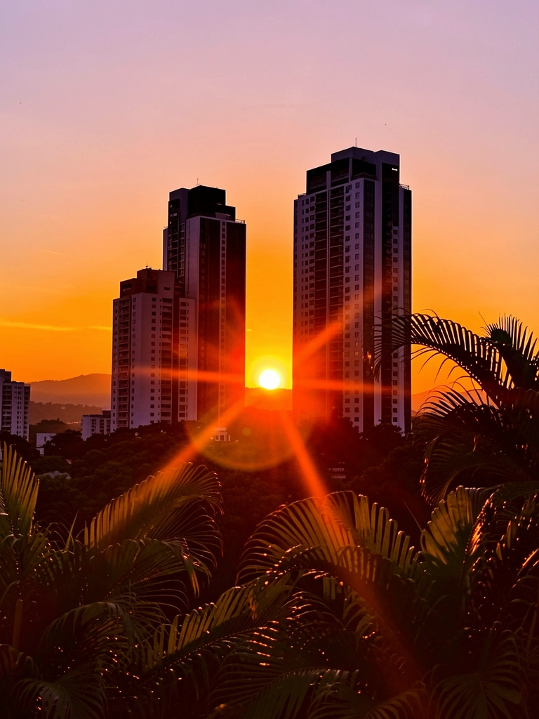 Arranha-céus escuros contra pôr do sol laranja com flare, vegetação tropical dourada ao fundo.
