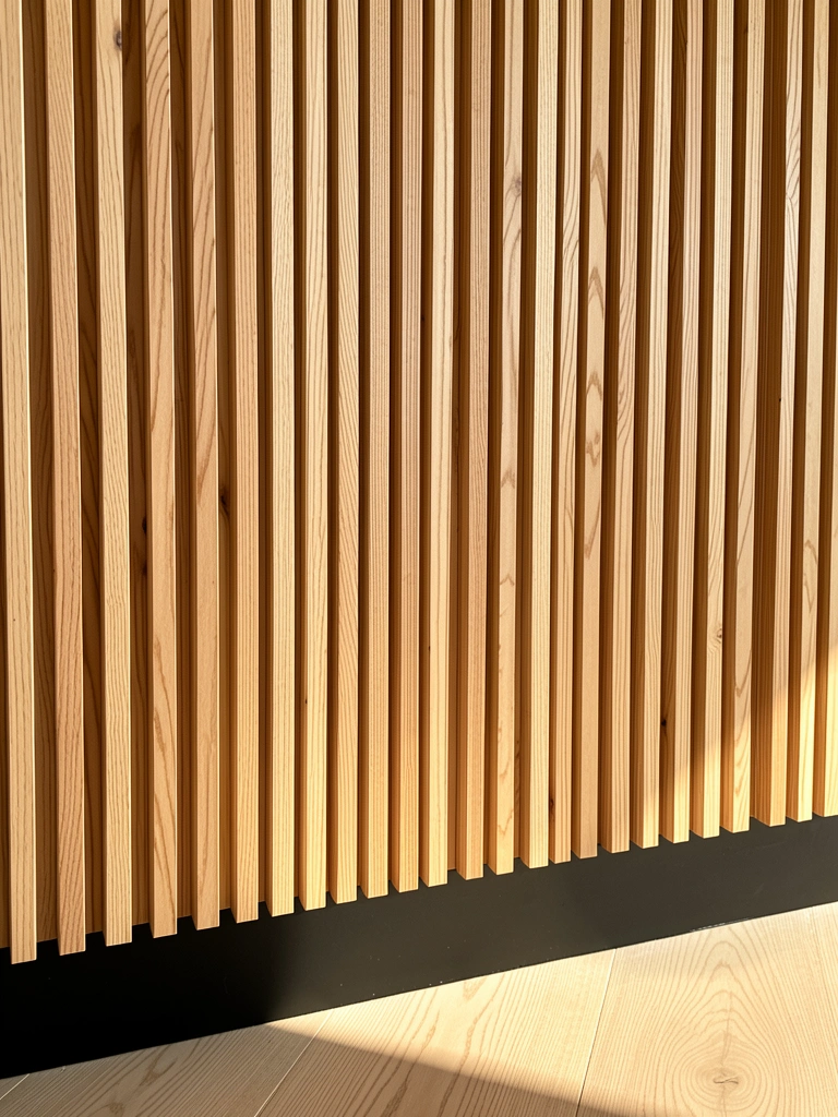 Painel ripado de madeira clara com base preta fosca, sombra projetada no assoalho de madeira clara, luz natural quente.