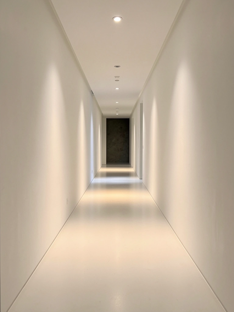 Corredor branco com iluminação embutida, piso polido e paredes lisas, luz refletida no chão cria sombras fortes e perspectiva alongada.