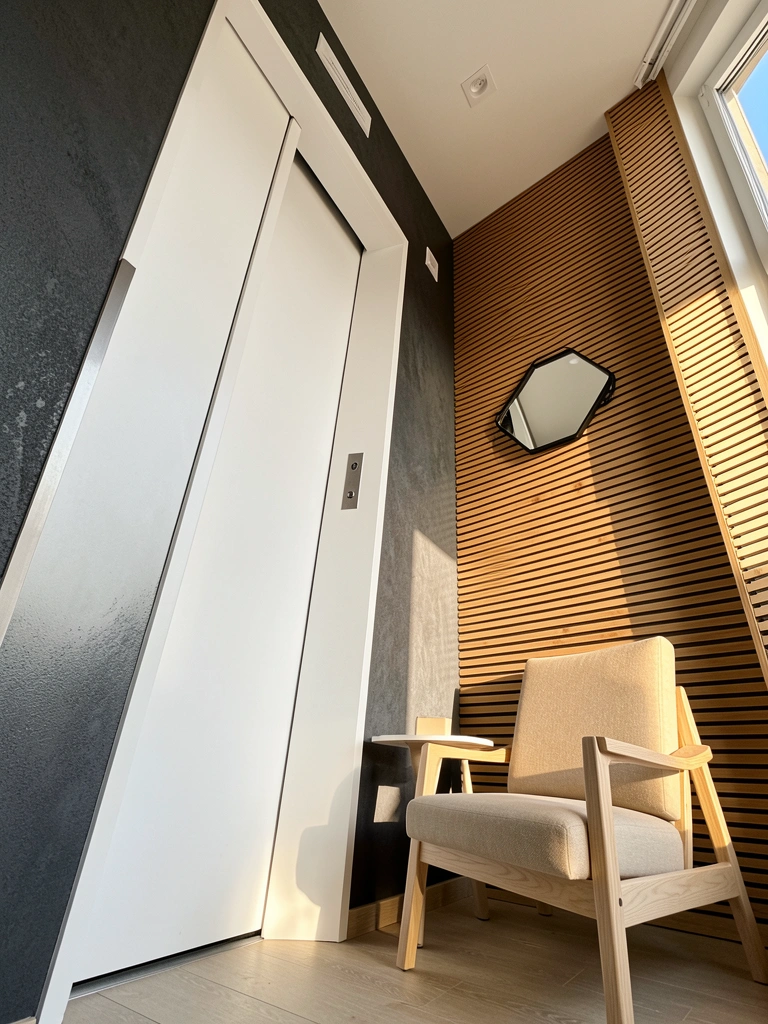 Parede texturizada cinza escuro, elevador branco e poltrona de estrutura clara com mesa lateral, luz natural e espelho inclinado ampliam a profundidade.