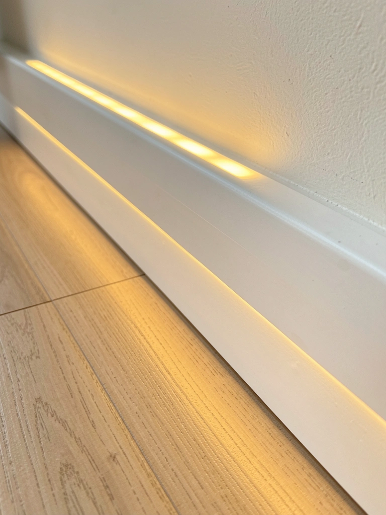Rodapé branco com iluminação LED embutida, brilho dourado no piso de madeira clara, luz solar difusa e sombra projetada.